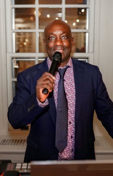 A man in a suit and tie speaks into a microphone indoors, standing in front of large windows at night.